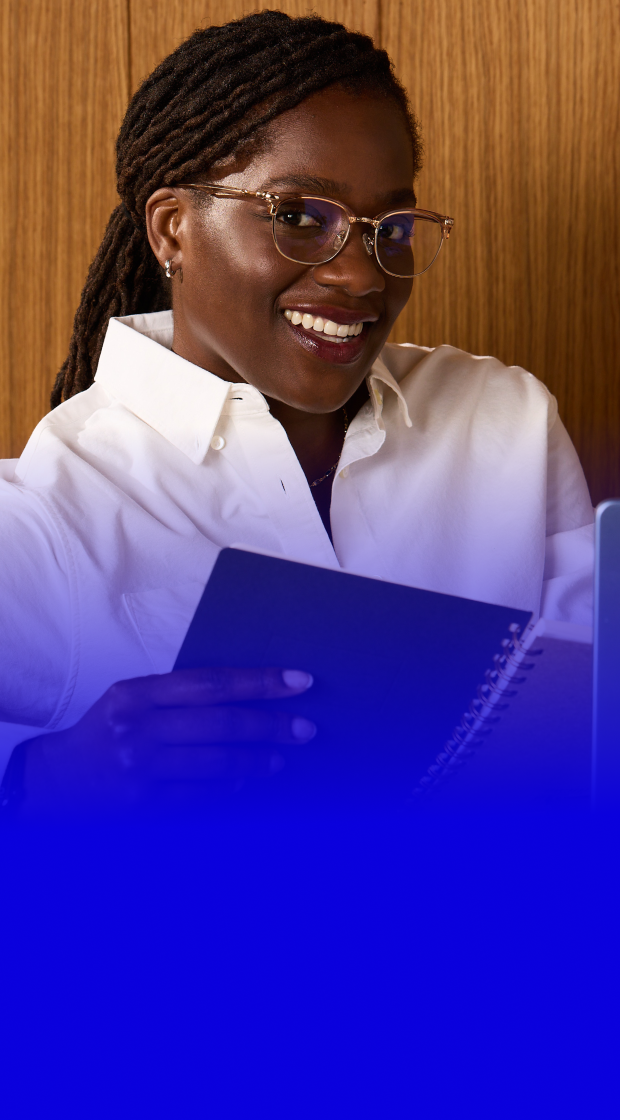 Close-up of a smiling woman with braided dark hair wearing clear round eyeglasses and a white button-down shirt, sitting in front of a wooden wall.