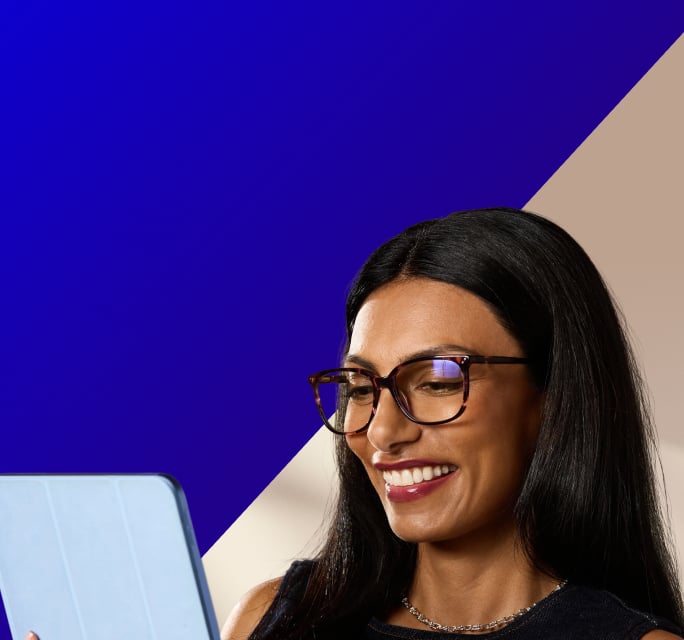 Smiling woman with long dark hair wearing tortoiseshell eyeglasses while looking down at a light blue tablet, set against a split background of deep royal blue and beige.