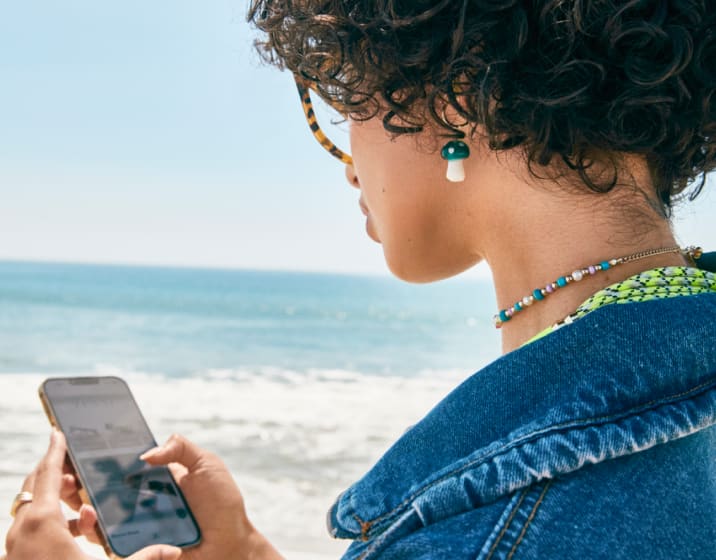 A woman is using her Blokz blue light glasses to look at her phone while on the beach.