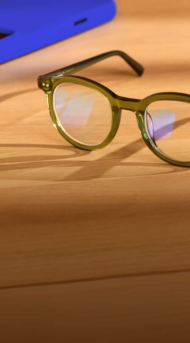 Translucent orange round eyeglasses resting on a wooden side table next to a blue smartphone.