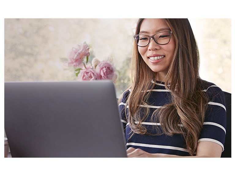 A young woman with long brown hair wearing Blokz by Zenni blue blocker glasses smiles while using her laptop computer.