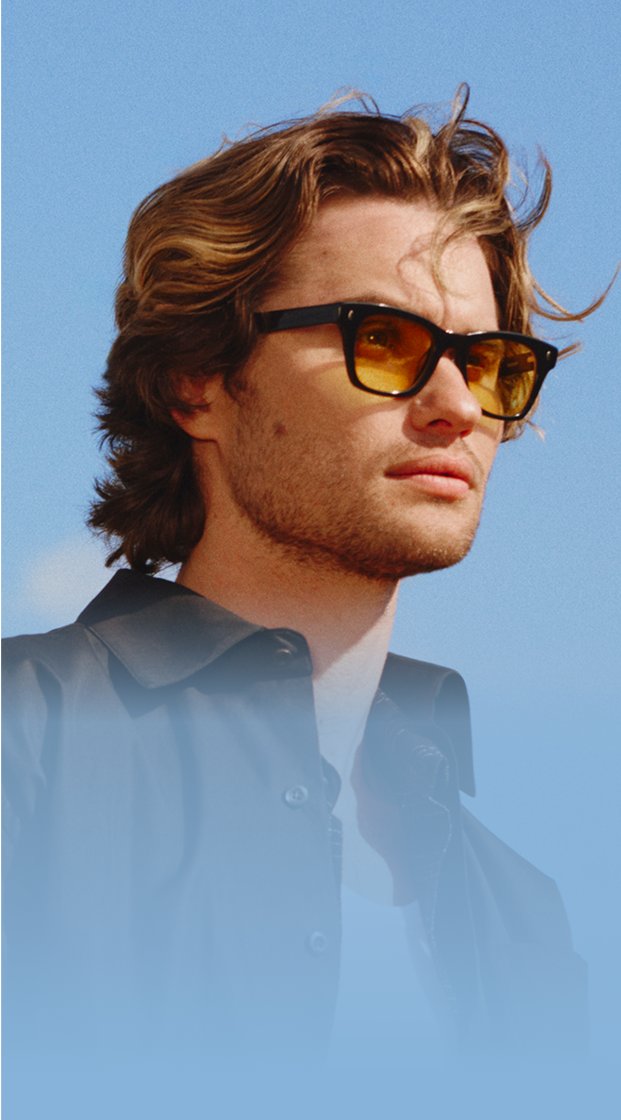 Chase Stokes wearing Zenni black frames with tinted lenses, standing outside with a clear blue sky in the background.