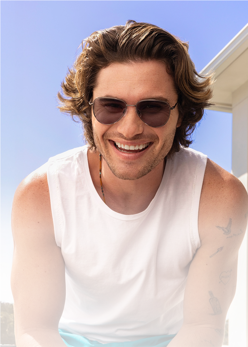 Chase Stokes wearing sunglasses leans forward in a white tank top under a clear blue sky.