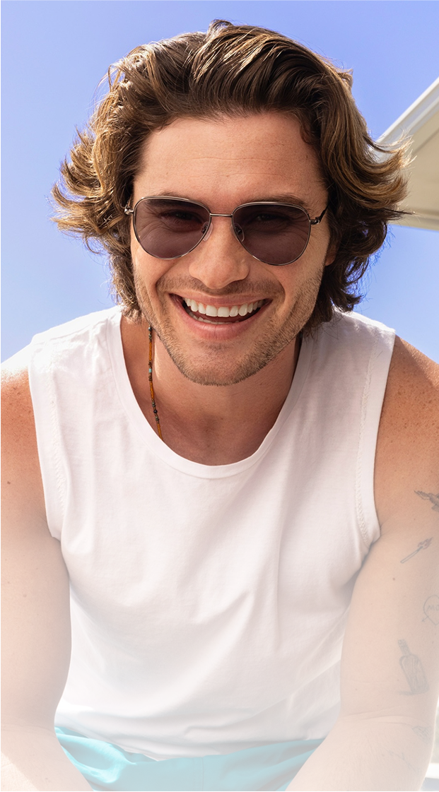 Chase Stokes wearing sunglasses leans forward in a white tank top under a clear blue sky.