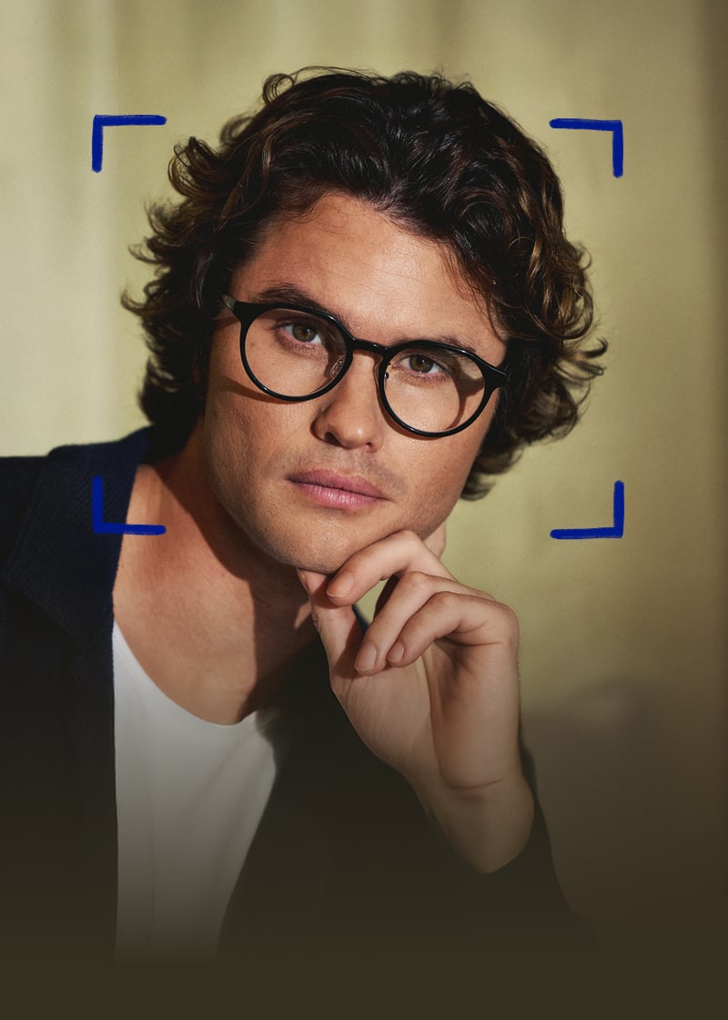 Chase Stokes is wearing round black Zenni glasses in front of a gold curtain backdrop.