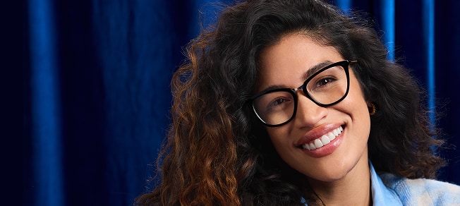 Woman wearing Zenni black square glasses and a light blue shirt, smiling against a deep blue curtain.