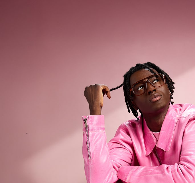 Man in pink jacket looking up while wearing Zenni oversized brown square glasses, pink background.