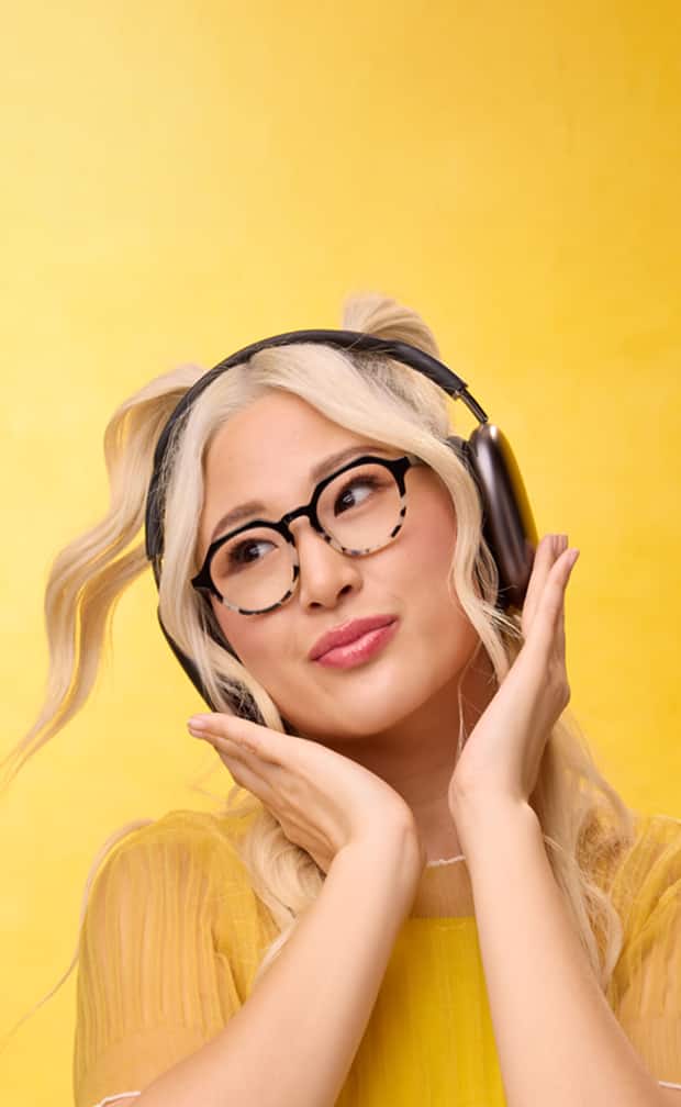 Blonde woman wearing round Zenni glasses, smiling with headphones on against a yellow background.