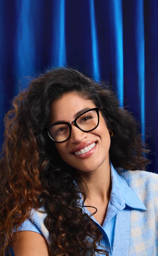 Woman wearing Zenni black square glasses and a light blue shirt, smiling against a deep blue curtain.