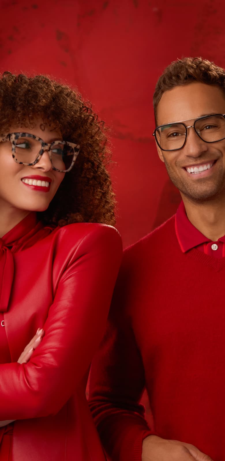 Two models in bold Zenni glasses posing against a rich red background - one woman with voluminous curls and a man in a red jacket.