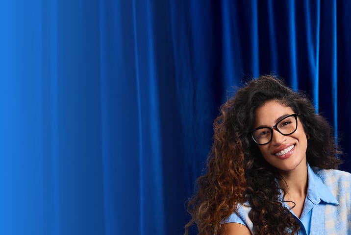 Woman wearing Zenni black square glasses and a light blue shirt, smiling against a deep blue curtain.