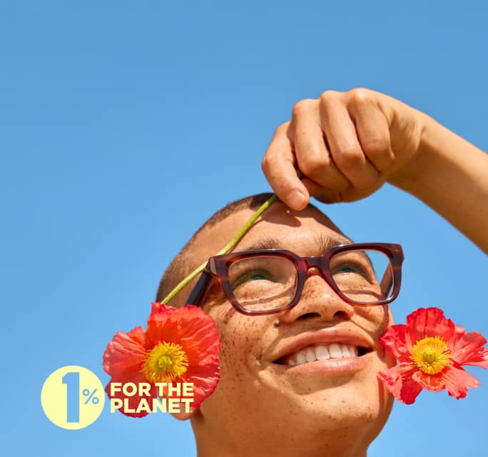 A smiling person with freckles wearing thick, square tortoiseshell eyeglasses, holding a red poppy flower over their eye against a clear blue sky, featuring a '1% FOR THE PLANET' logo.
