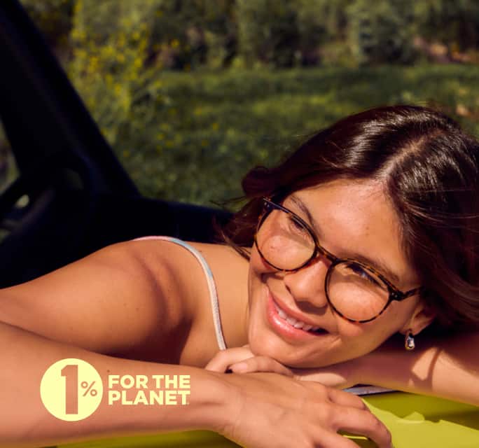 A woman with medium brown hair and tortoiseshell glasses smiles while leaning on a bright yellow surface, with green foliage in the softly blurred background on a sunny day.