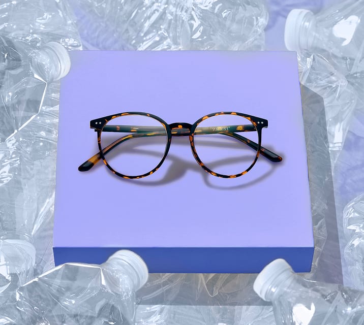 Tortoiseshell EcoBloomz eyeglasses displayed on a purple platform surrounded by recycled plastic, representing the eco-friendly eyewear collection.