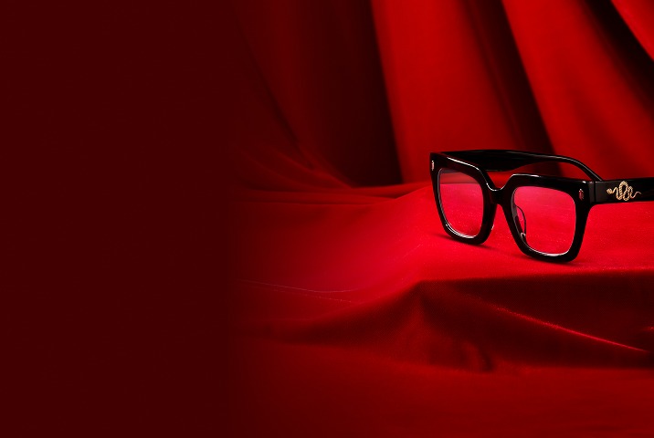 A pair of thick black rectangular glasses featuring a gold snake engraving on the temple, resting on a draped red velvet background.