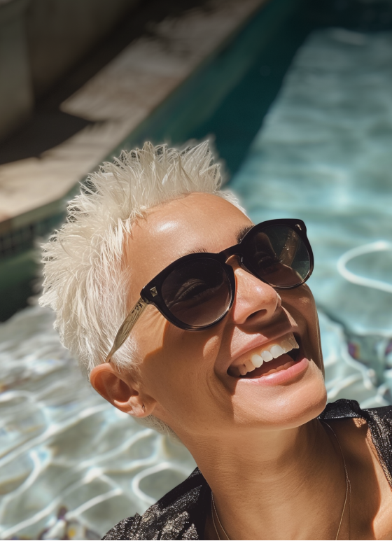 Close-up of a person with short white-blonde hair and stylish frames, facing the sun by a swimming pool, highlighting sun protection.