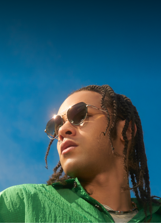 A fashion-forward individual with braids wearing gold aviator EyeQLenz frames from Zenni, gazing pensively into the distance under the sunlight.