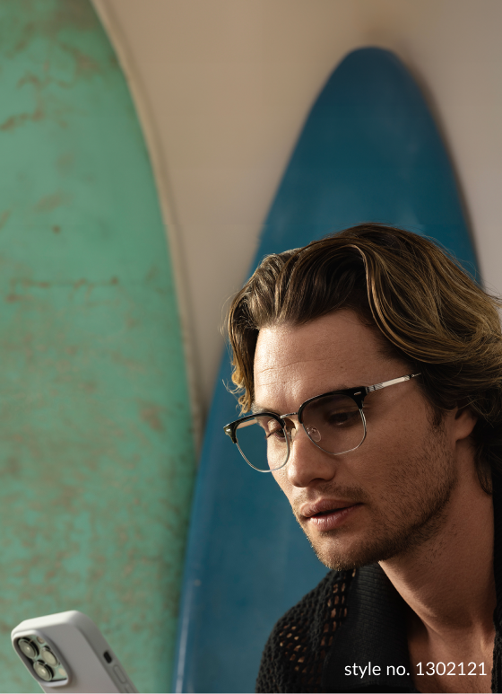 Chase Stokes checking his phone, wearing Zenni EyeQLenz glasses and a black shirt, with a surfboard in the background.