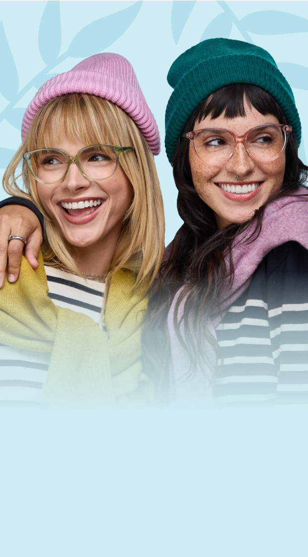 A blond woman wearing clear green glasses and a pink knit hat being hugged by a brunette woman wearing clear pink glasses and a green knit, on a light blue background with a foliage motif.