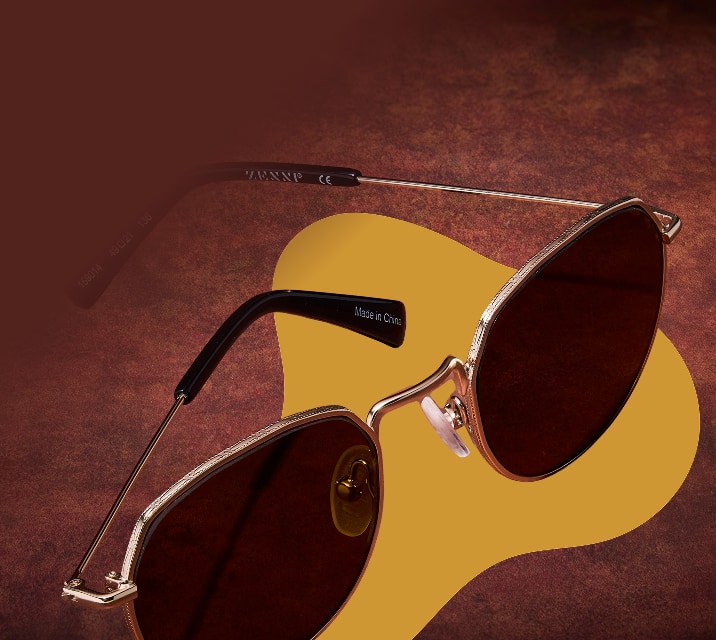 Gold wireframe sunglasses with dark brown tinted lenses and textured rims floating over a mustard yellow shape against a warm brown background.