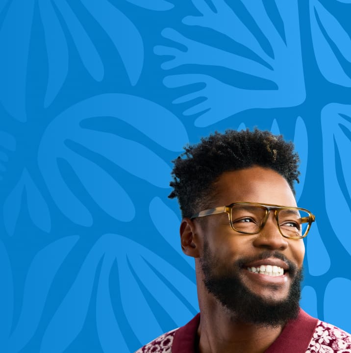 Smiling man wearing tortoiseshell eyeglasses and a patterned red cardigan, standing in front of a blue background with abstract shapes.