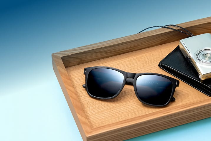 Matte black sunglasses with dark lenses resting on a rectangular wooden tray next to a black leather wallet and a silver compact camera, set against a blue gradient background.