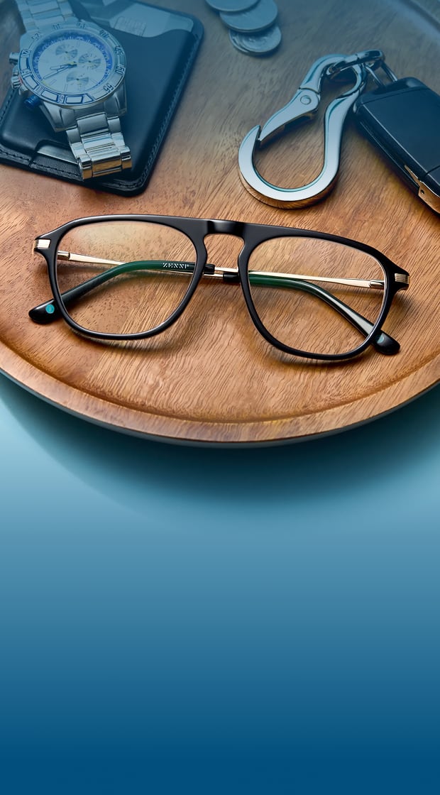 Black aviator-style eyeglasses with gold metal temples resting on a round wooden tray alongside a silver watch, leather wallet, coins, and a keychain with a carabiner.