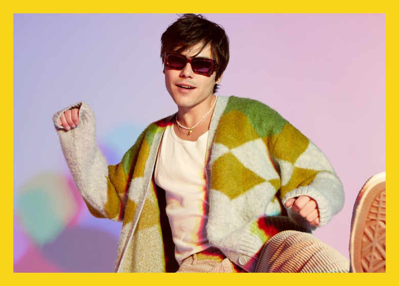 A person with tousled hair wearing Zenni sunglasses and a cozy, patterned cardigan over a white tee, exuding a cool festival vibe against a soft pastel backdrop.