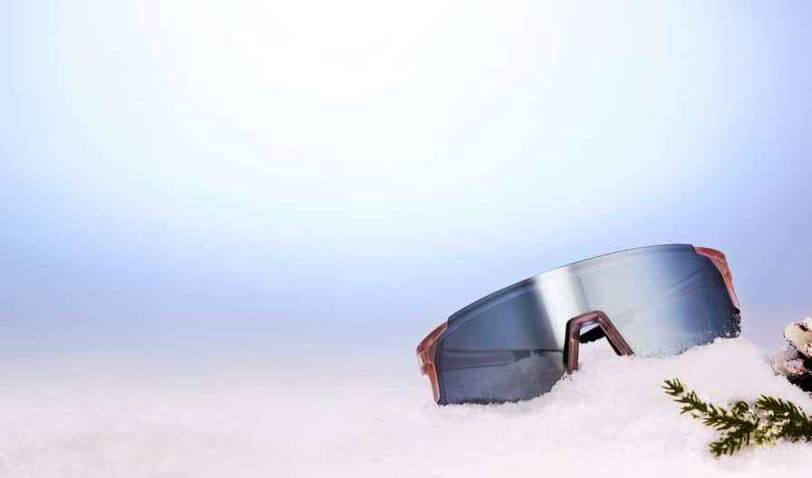 Zenni lleva Lentes de Sol con lentes espejadas sobre un montículo de nieve junto a una piña y una ramita perenne.