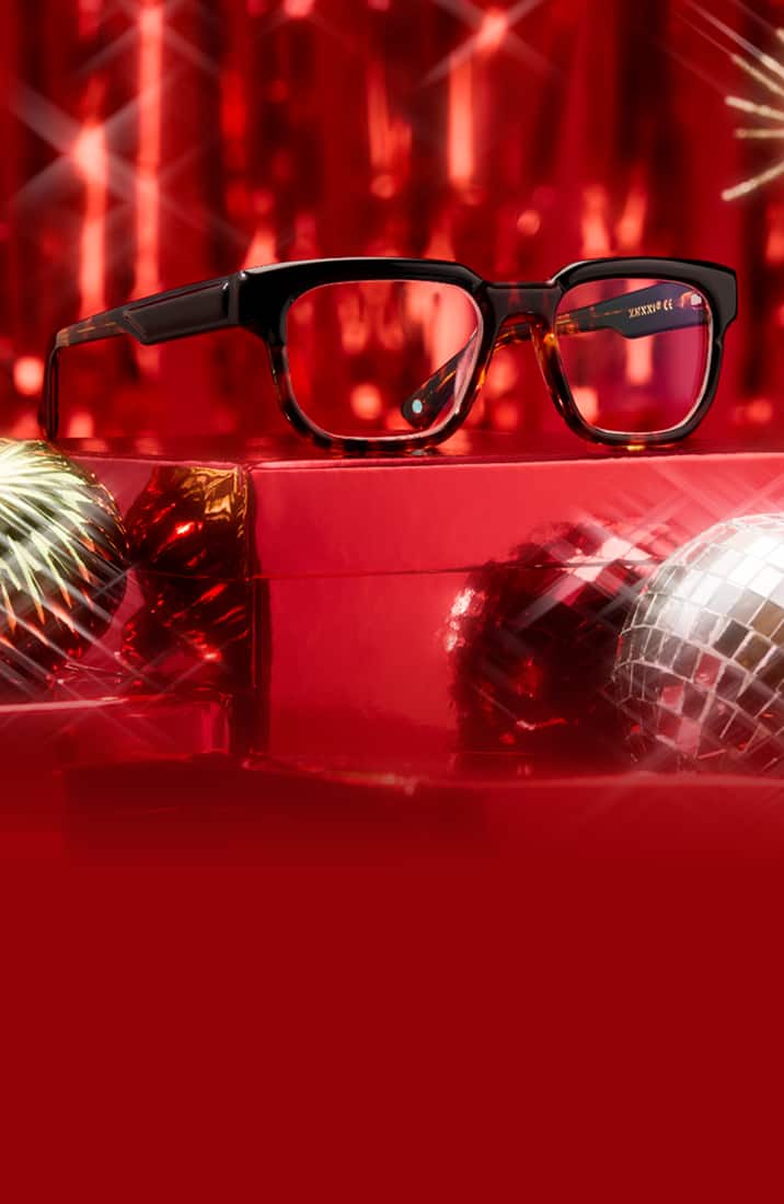 Stylish Zenni eyeglasses displayed with festive winter party props on a red foil tinsel background.