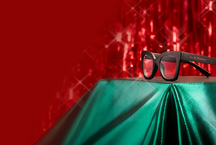 Black Zenni eyeglasses with "B & K Forever" custom engraving on temple arm against festive red and green holiday lights background.