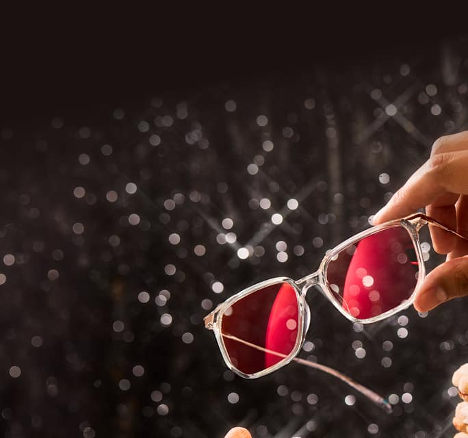 EyeQLenz with Zenni ID Guard eyeglasses with pink-tinted lenses held against sparkling holiday lights background.