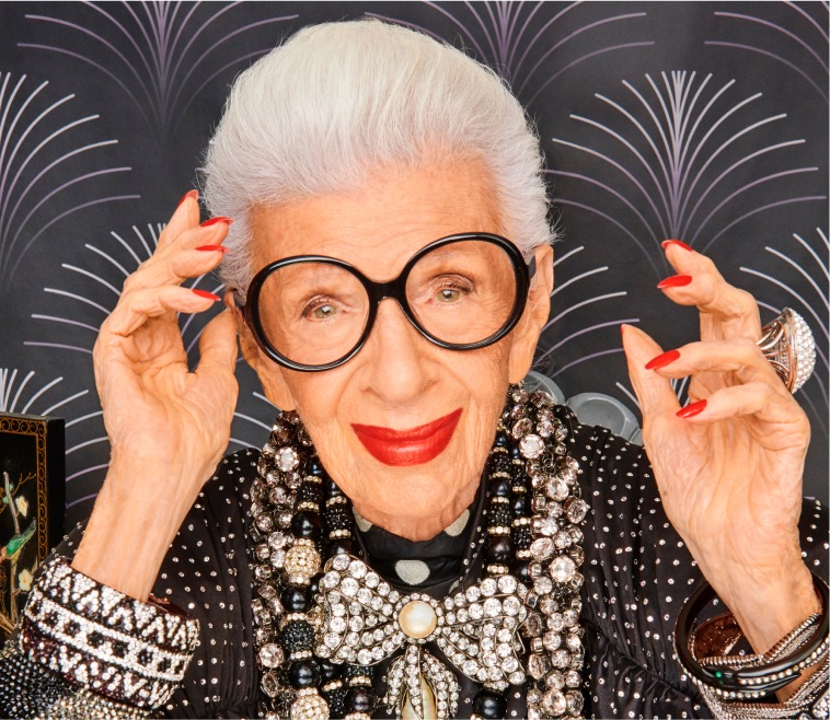 Image of Iris Apfel wearing Zenni Iris Apfel round glasses #4452521. She is wearing bright red lipstick and is in front of a black patterned wall.