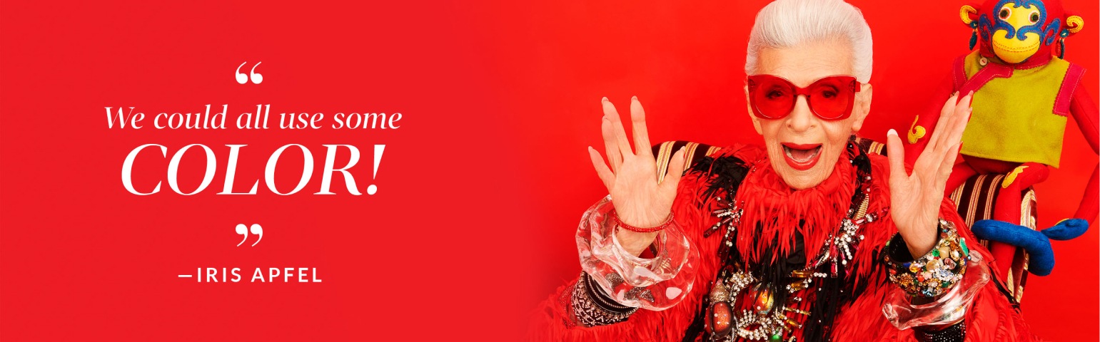 ‘We could all use some color!’ -Iris Apfel. Image of Iris Apfel wearing Zenni premium cat-eye sunglasses #1117918 against a red background, with a yellow toy monkey behind her.