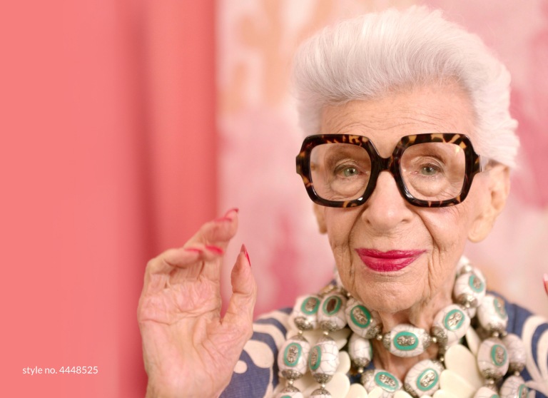 Watch Iris Apfel behind the scenes. Image of Iris Apfel wearing Zenni square glasses #4448525 standing in front of a pink background. Link to video.