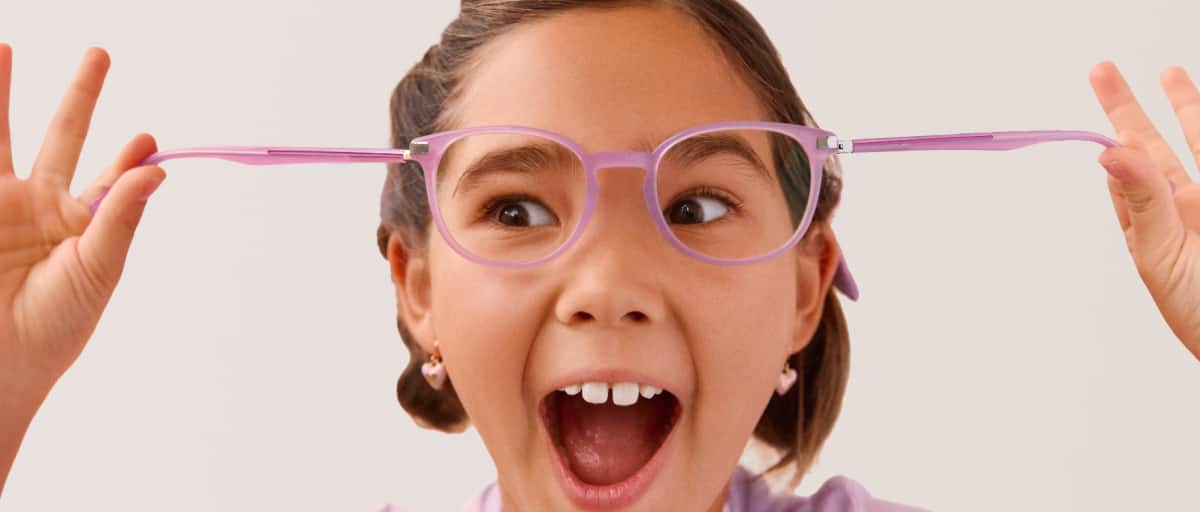 Close-up of an excited, wide-eyed girl with her mouth open in a happy shout, holding her translucent pink rectangular eyeglasses up to her face with both hands against a plain white background.