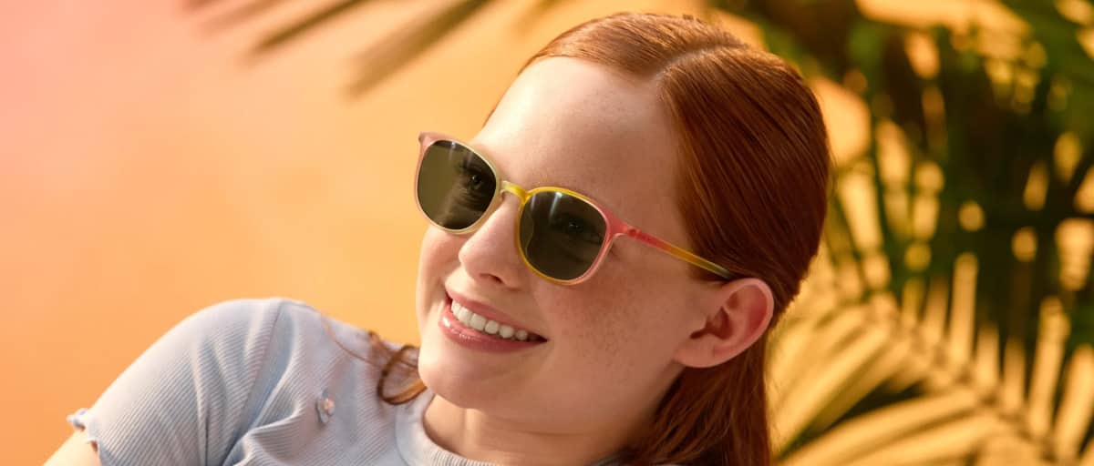 Close-up of a smiling girl with red hair looking over her shoulder, wearing round sunglasses with green-tinted lenses against a warm orange background with green palm leaves.