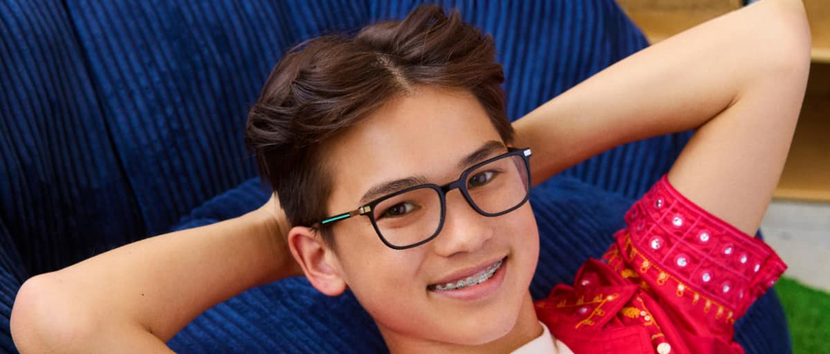 Close-up of a smiling boy with dark hair leaning back with his hands behind his head, wearing dark rectangular eyeglasses with green temples against a dark blue background.