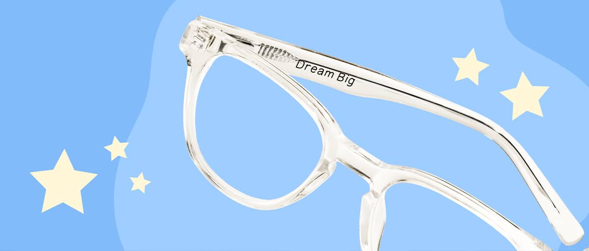 Clear transparent eyeglasses displaying the inscription Dream Big on the inside temple arm, set against a two-tone periwinkle blue graphic background decorated with pale yellow stars.