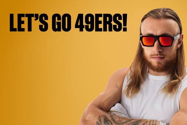 George Kittle, 49er, with long hair, wearing yellow lens sunglasses and white tank, orange background.