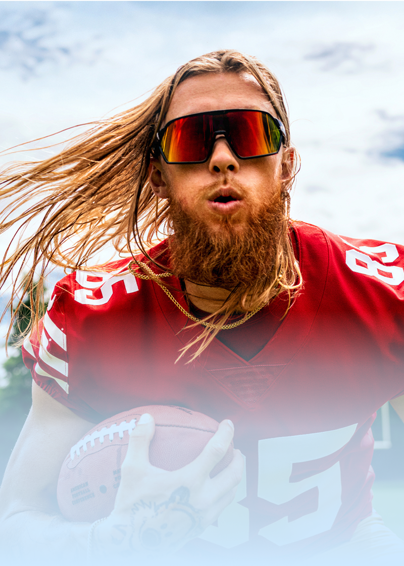 George Kittle in a red jersey and mirrored sport sunglasses, running on a football field while holding a football.