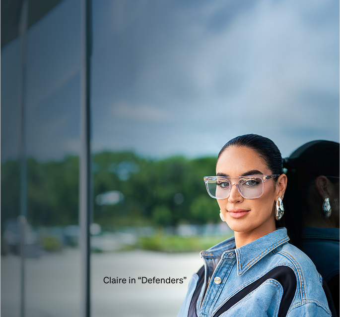 Claire wearing ‘Defenders’ - oversized transparent glasses, in a denim jacket, standing by a glass window, with a blurred green background.
