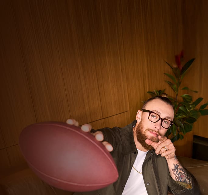 George Kittle wearing thick-rimmed tortoiseshell glasses, pointing toward the camera while holding a football in a room with wood-paneled walls.