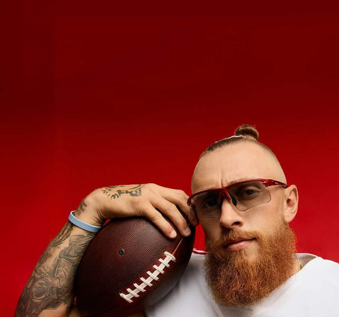 George Kittle sosteniendo un balón de fútbol americano, sobre un fondo rojo, usando lentes de sol Zenni rojos de la colección The Kittles.