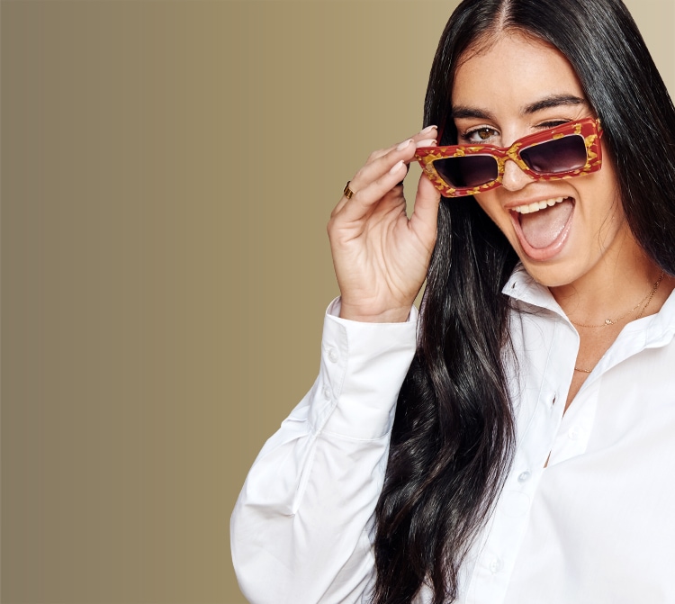 Claire Kittle, holding Zenni cat-eye glasses #4449318 on her nose and smiling, in front of a gold background.