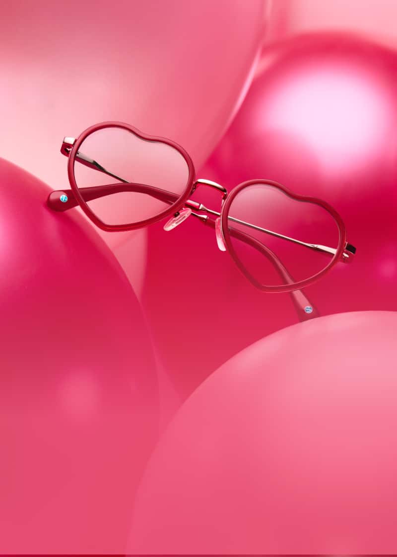 A pair of gold-rimmed heart-shaped glasses with red arms leaning against a group of shiny pink balloons.