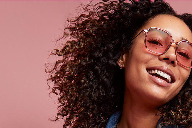 Woman with curly hair smiling, wearing a denim jumpsuit and Zenni FL-41 rose-tinted glasses, against a pink background.