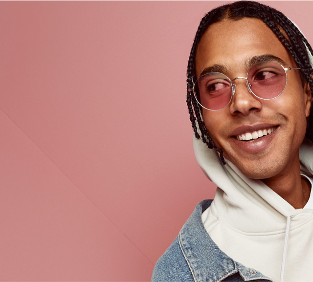 Stylish man with braids wearing a white hoodie, denim jacket, and Zenni FL-41 rose-tinted glasses, posing with a smile against a pink background.
