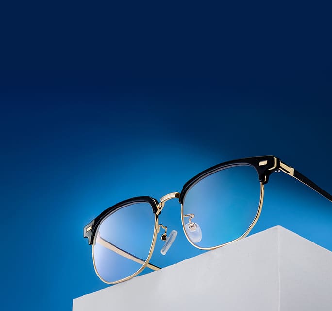 A pair of black and gold browline glasses resting on a white geometric block against a vibrant blue gradient background.