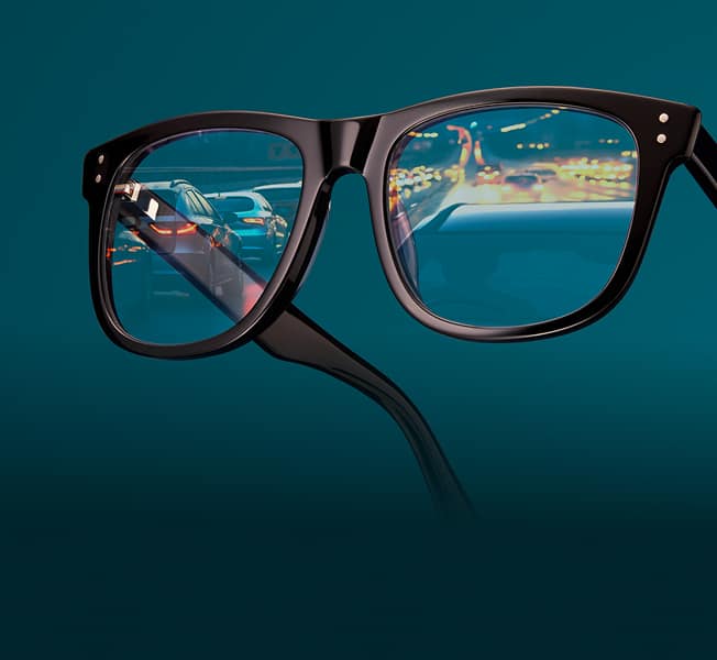 Square black glasses with nearly clear night driving lenses, reflecting a street at night. Background is dark teal.